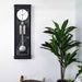 Black wall clock with pendulum next to a potted plant on a white wall