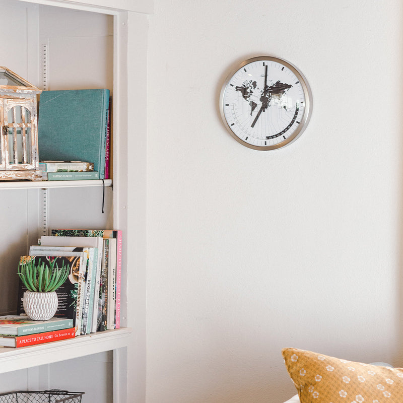 World map clock hanging on white well near bookshelf with colorful books and small plant. 