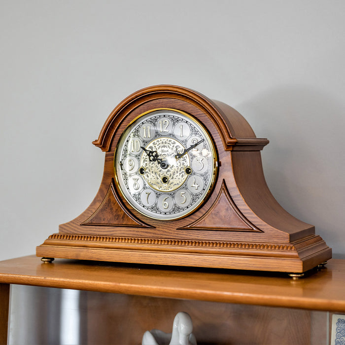 Wooden mantle clock with intricate face on shelf with white wall behind