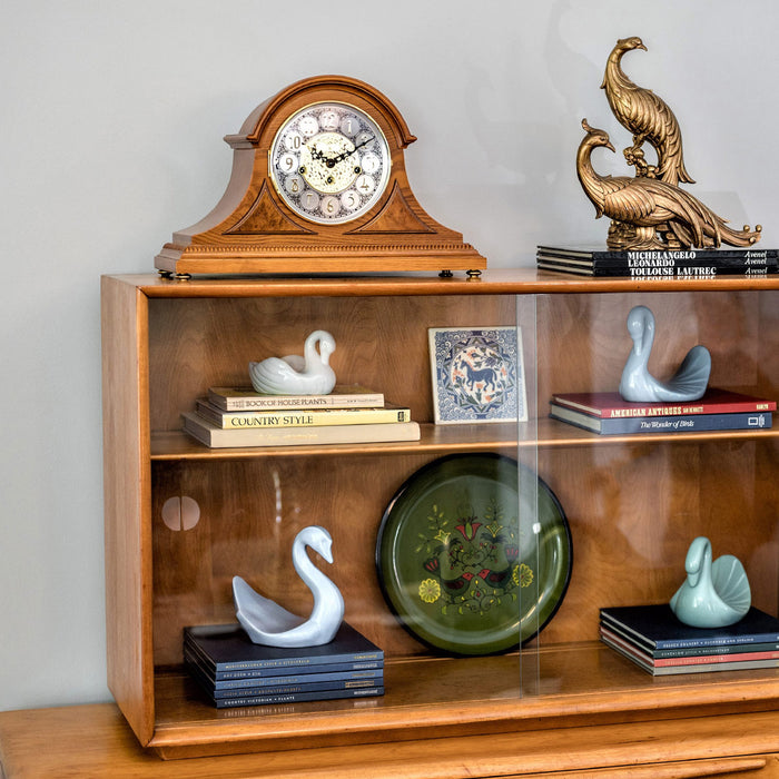 Wooden Mantle clock with intricate face on shelf with books and knick-knacks in room with white walls 