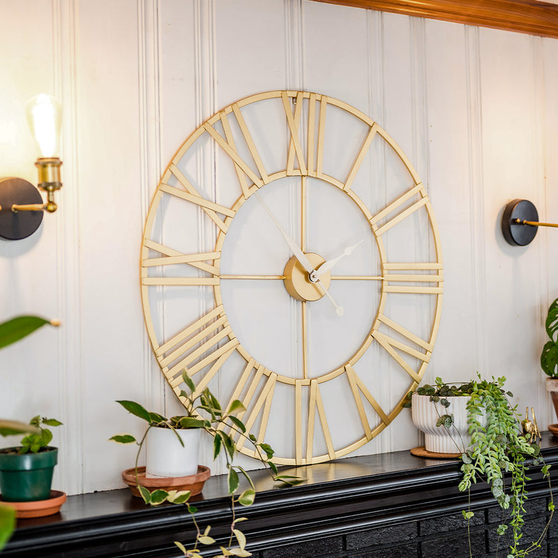 Roman numeral clock with airy design on black shelf flanked by green plants in room with white walls.