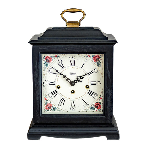 Black decorative clock with floral design and holes on the face for winding on a white background