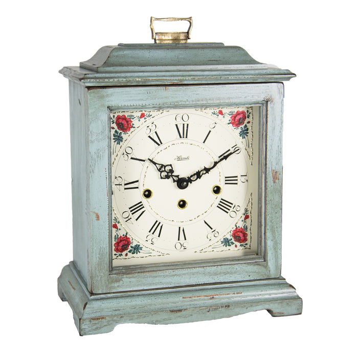 Blue decorative clock with floral design and holes on the face for winding on a white background