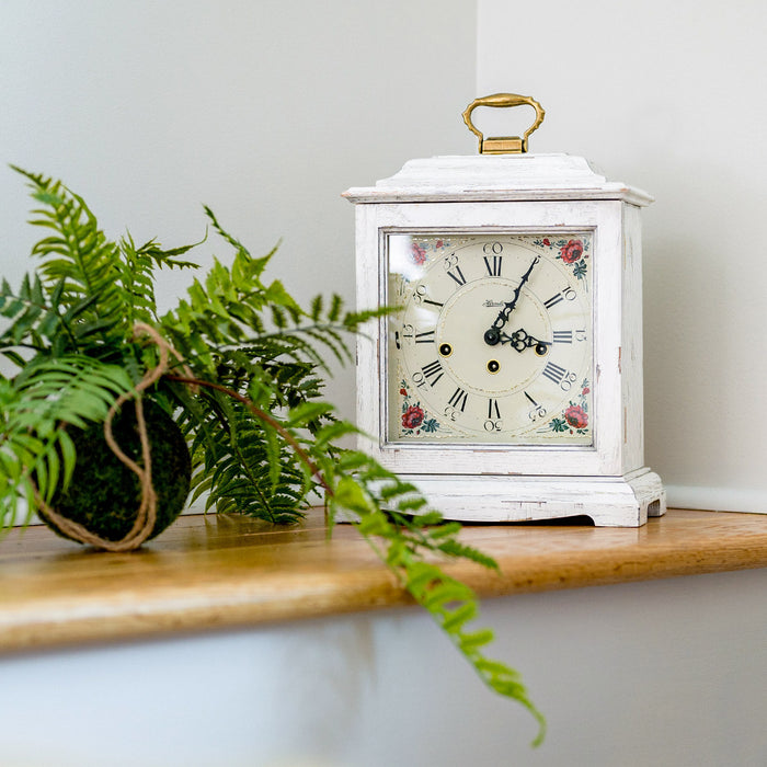 White vintage-style clock with floral design on a wooden surface next to a green plant.