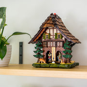 Decorative weather station with green shutters and deer figurines beneath pine trees sitting on shelf next to green plant.