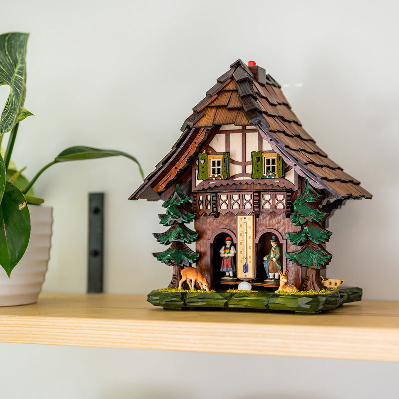 Decorative weather station with green shutters and deer figurines beneath pine trees sitting on shelf next to green plant.