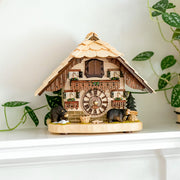 Wooden cuckoo clock with bear figures on white shelf with green plant nearby.