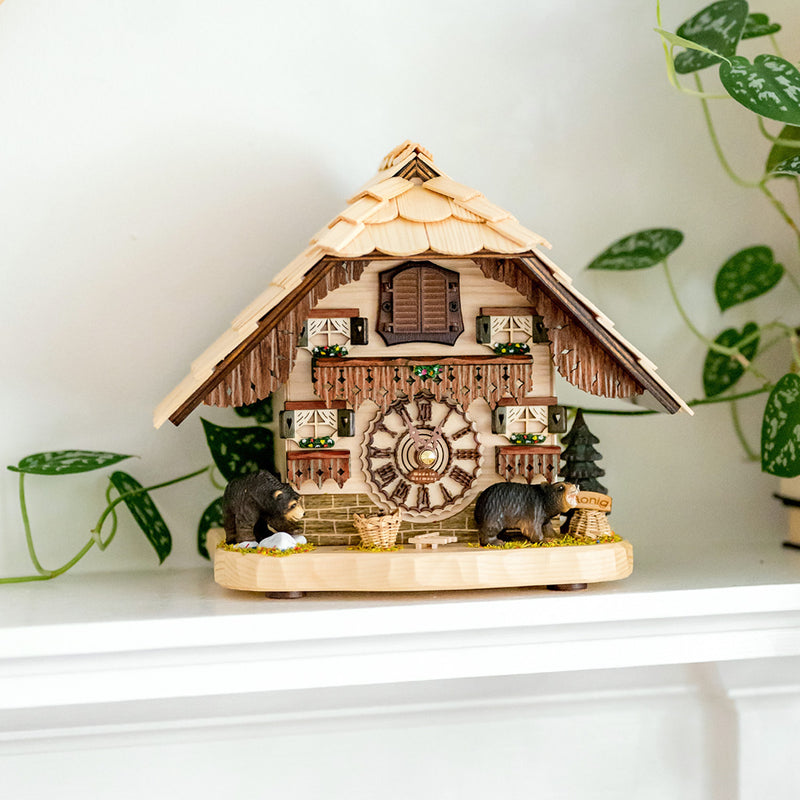 Wooden cuckoo clock with bear figures on white shelf with green plant nearby.