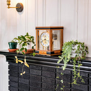 Light cherry mantel clock with glass sides on black brick mantel surrounded by green plants.