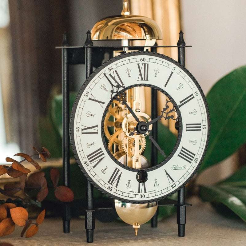 Up close roman numeral face on decorative mantel clock with visible brass gears on table with green plants.