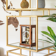 Walnut wood mantel clock on glass and metal shelf near green plants.