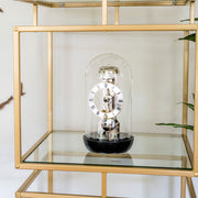 Black mantel clock with glass dome on gold and glass shelf in front of white wall.