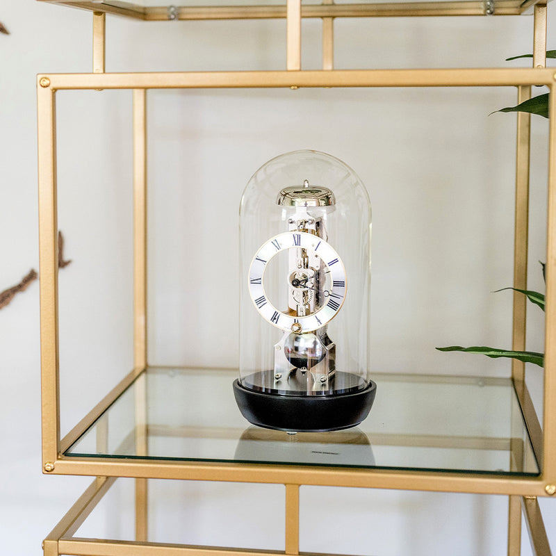 Black mantel clock with glass dome on gold and glass shelf in front of white wall.