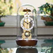 Walnut wood mantel clock with glass dome on reflective surface with green plants in background.