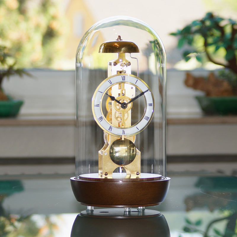 Walnut wood mantel clock with glass dome on reflective surface with green plants in background.