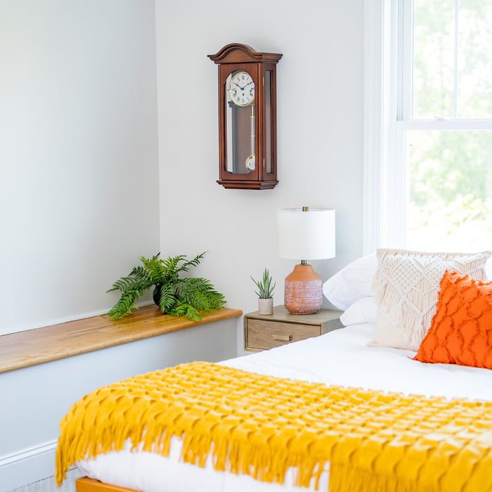 Cherry wood wall clock hanging above nightstand with lamp next to bed with yellow bedspread 