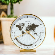 Brass world map clock on white countertop in front of green plants. 