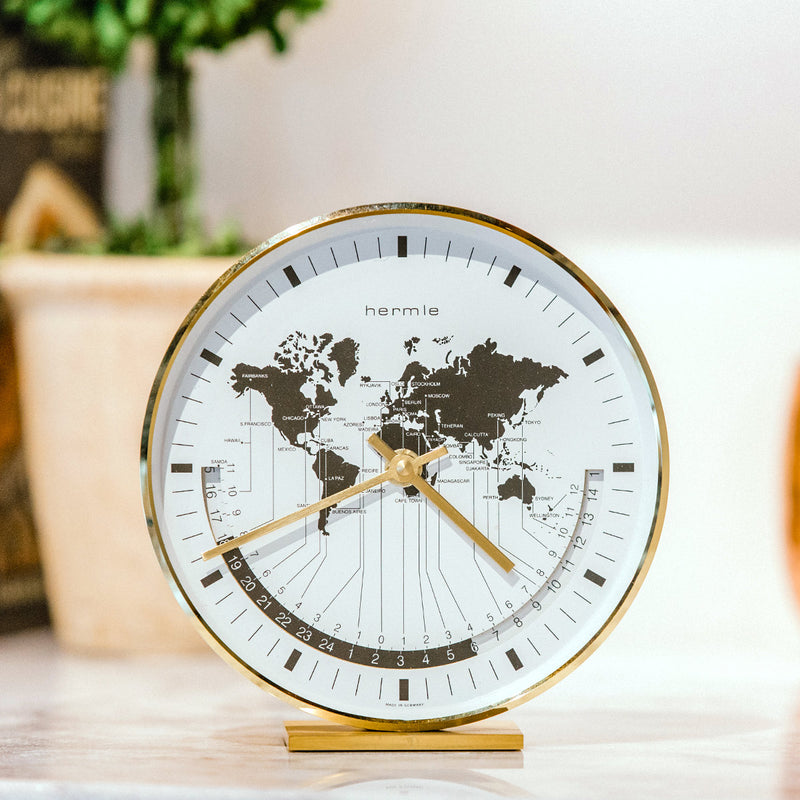 Brass world map clock on white countertop in front of green plants. 