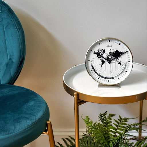 Nickel world map clock on gold and white side table next to blue chair.