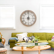 Wooden clock with Roman Numerals hanging on white wall above green sofa in living room.
