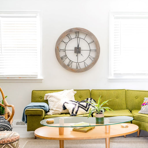Wooden clock with Roman Numerals hanging on white wall above green sofa in living room.