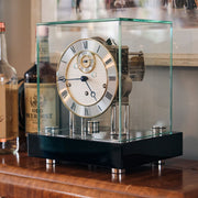 Black desk clock with glass case sitting on wooden shelf next to wine bottles