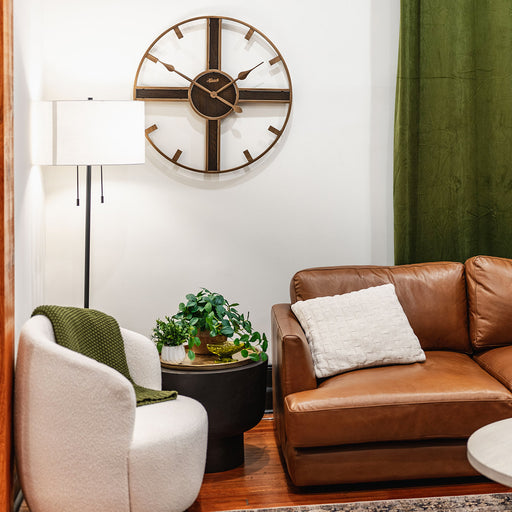 Rustic wood and metal clock hanging on white wall above leather couch next to green curtain.