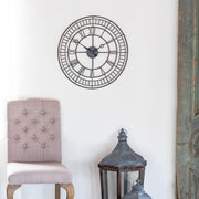 Large round metal wall clock with roman numerals hanging on white wall above beige chair and large decorative lanterns.