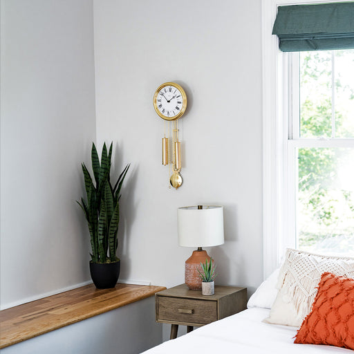 Ornate gold finish wall clock hanging on white wall above nightstand and lamp with green plants nearby.