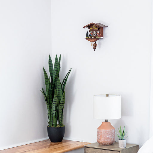 Wooden cuckoo clock with squirrel figurine hanging on white wall above tall green plant and decorative lamp.