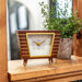 Retro style wooden clock on wood shelf with green plant behind.