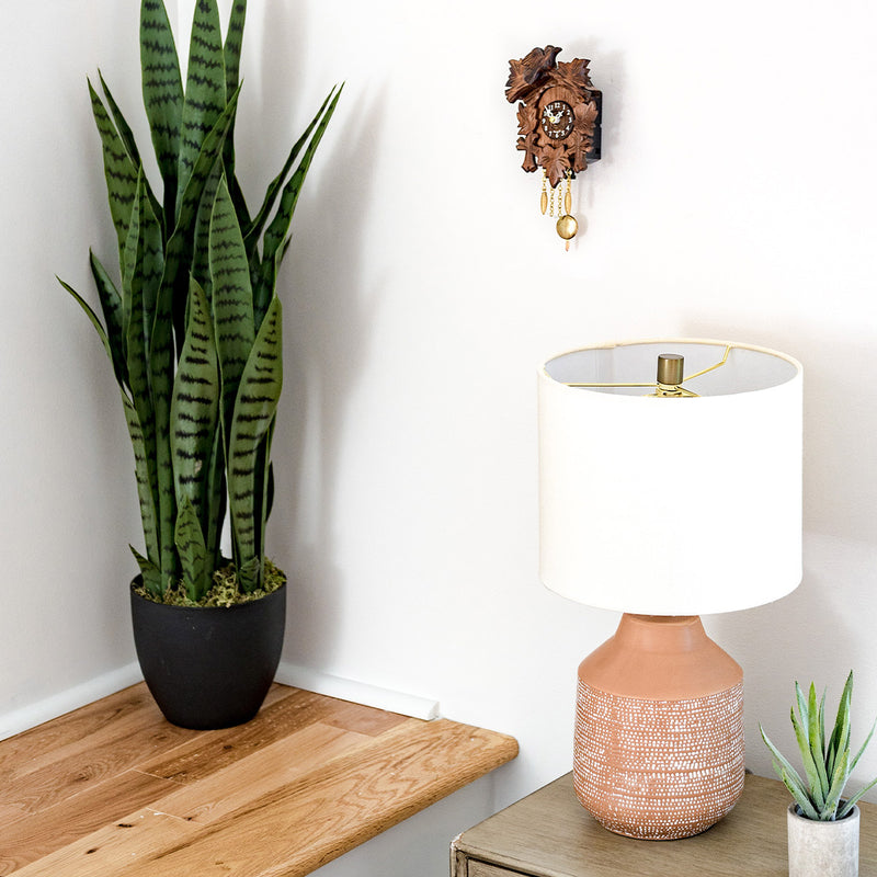 Small wooden cuckoo clock hanging on white wall above green plant and table lamp.