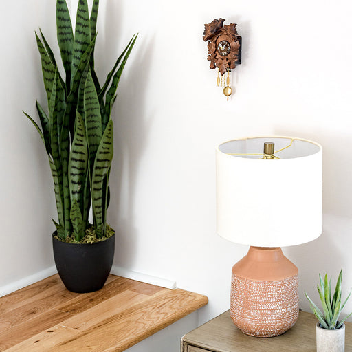 Small wooden cuckoo clock hanging on white wall above green plant and table lamp.