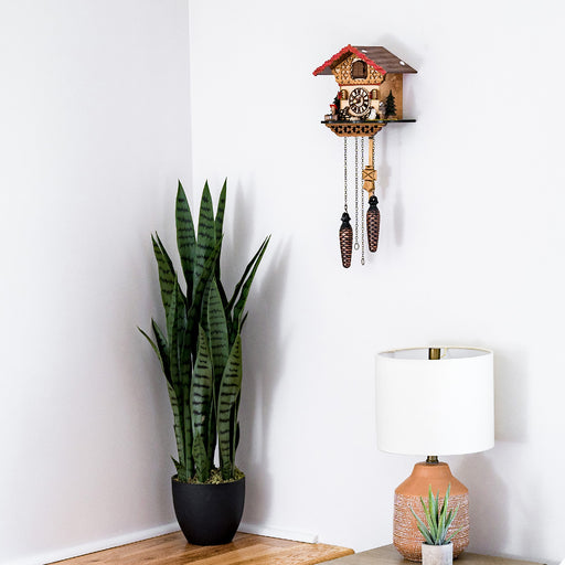 Decorative wooden cuckoo clock hanging on white wall above table lamp and tall potted plant 