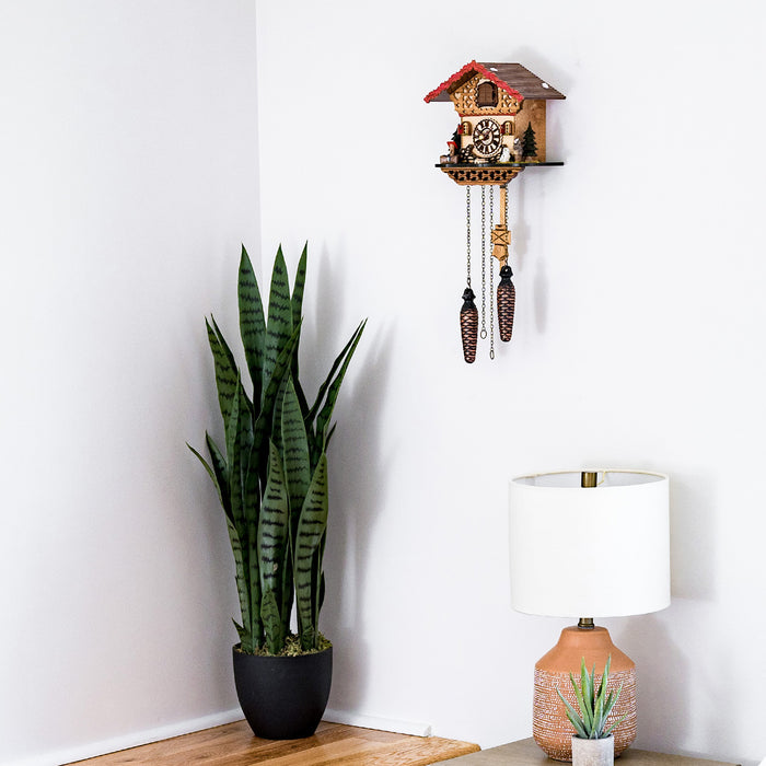 Decorative wooden cuckoo clock hanging on white wall above table lamp and tall potted plant 