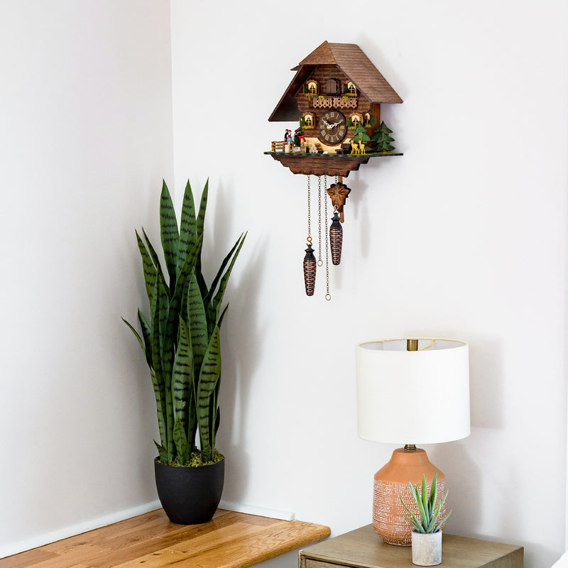 Chalet style cuckoo clock hanging on white wall above green plant and beige lamp.