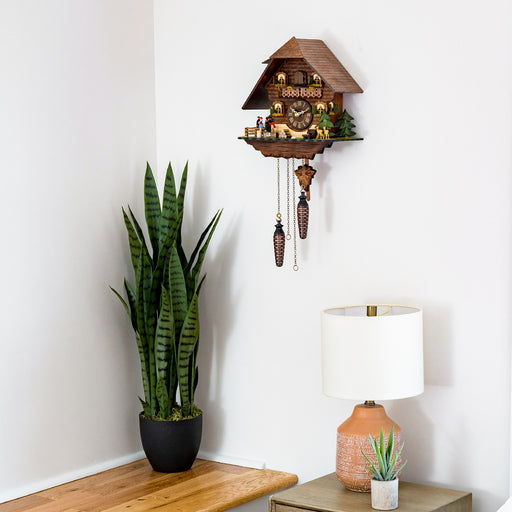 Chalet style cuckoo clock hanging on white wall above green plant and beige lamp.
