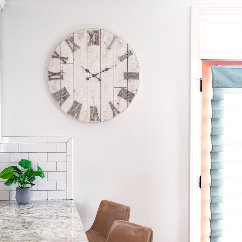 Rustic wood clock hanging on white kitchen wall above table with green plant and leather chairs. 