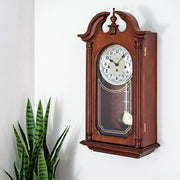 Left facing view of Hopewell wooden vintage style wall clock by Hermle on white wall with plant in corner.