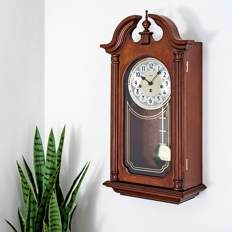 Left facing view of Hopewell wooden vintage style wall clock by Hermle on white wall with plant in corner.