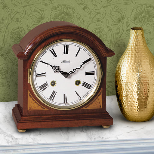 Wooden barrister style, vintage look,  mantel clock on marble mantel next to golden vase with floral pattern wall in background