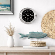 Ship's Bell clock by on a wall above a table with a fish sculpture and rope.