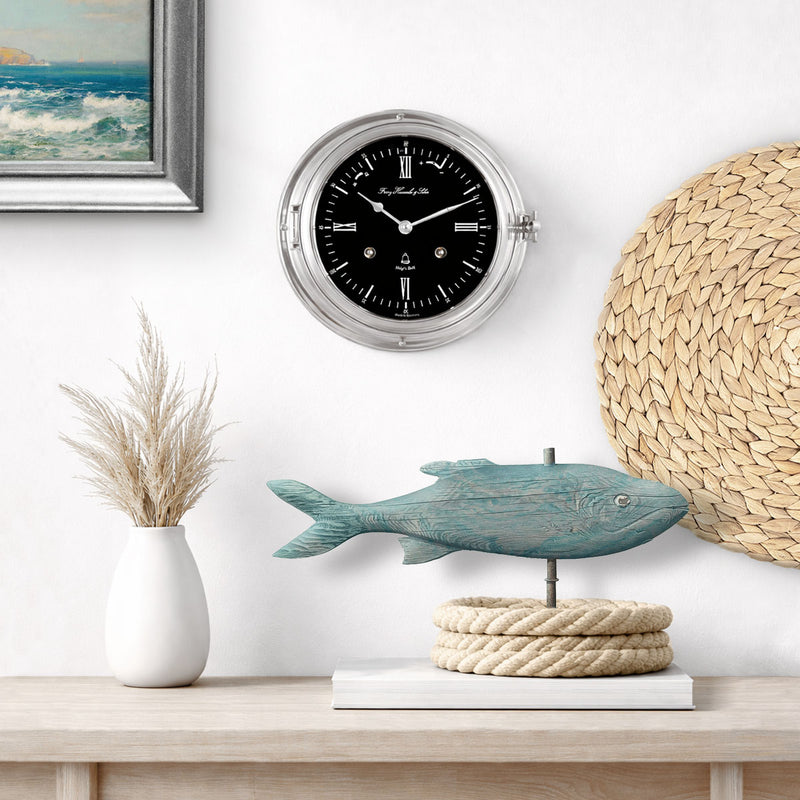 Ship's Bell clock by on a wall above a table with a fish sculpture and rope.