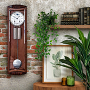 Wooden wall clock with pendulum on a brick wall with plants and books in the background