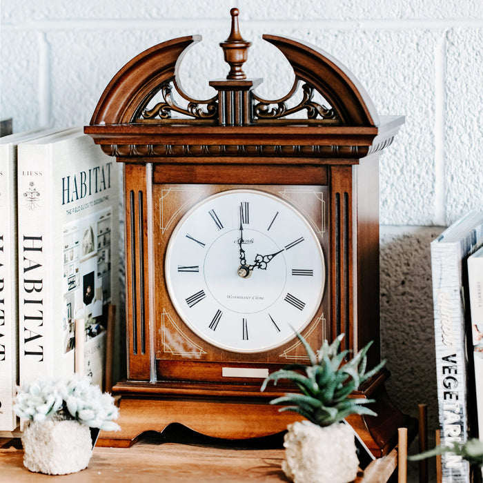 Wooden clock with decorative top on wooden dresser with books and plants.