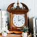 Wooden clock with decorative top on wooden dresser with books and plants.