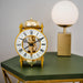 Brass mantel clock with visible intricate gears on green table in front of lamp