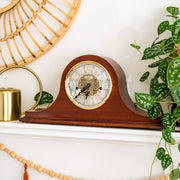 Cherry wood mantel clock on white mantel next to green plants. 