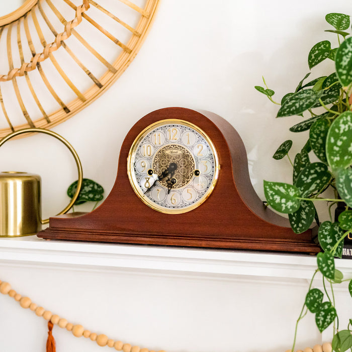 Cherry wood mantel clock on white mantel next to green plants. 