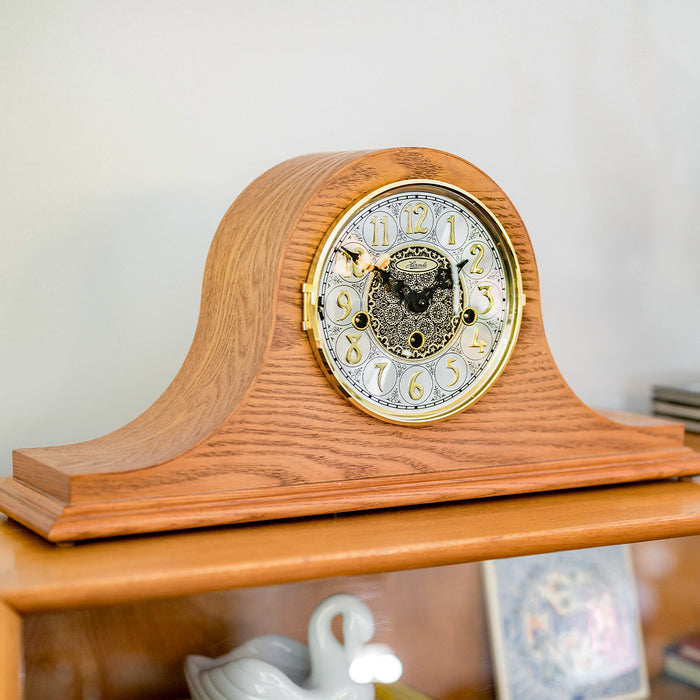 Light Oak mantel clock on wooden dresser in front of white wall. 